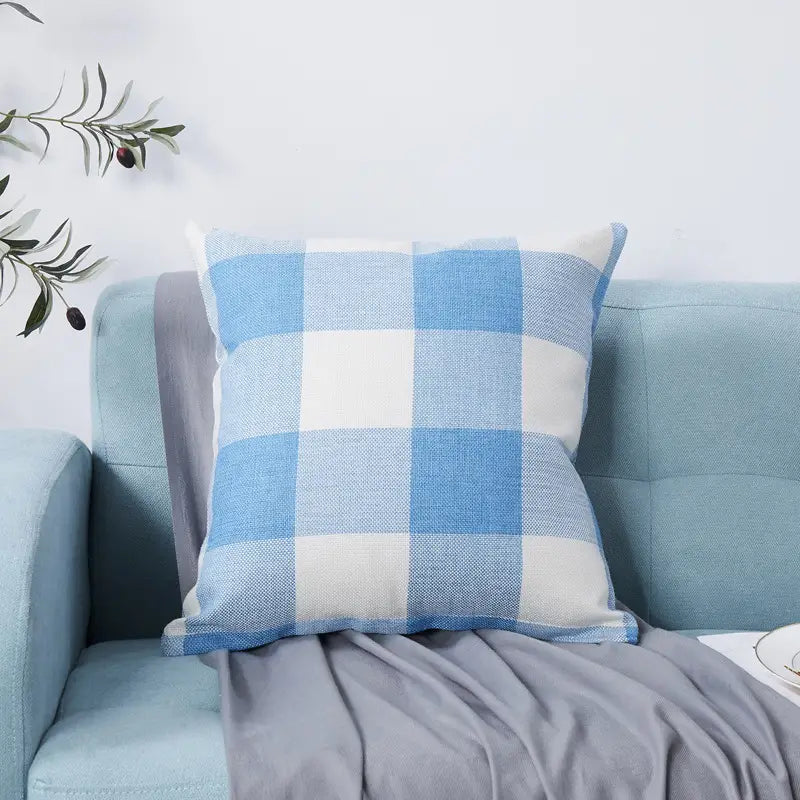 A square throw pillow featuring a large-scale gingham plaid pattern in soft light blue and white. The cover has a visible linen-like texture and is displayed on a light blue sofa with a grey blanket draped beside it, creating a bright and airy coastal aesthetic.