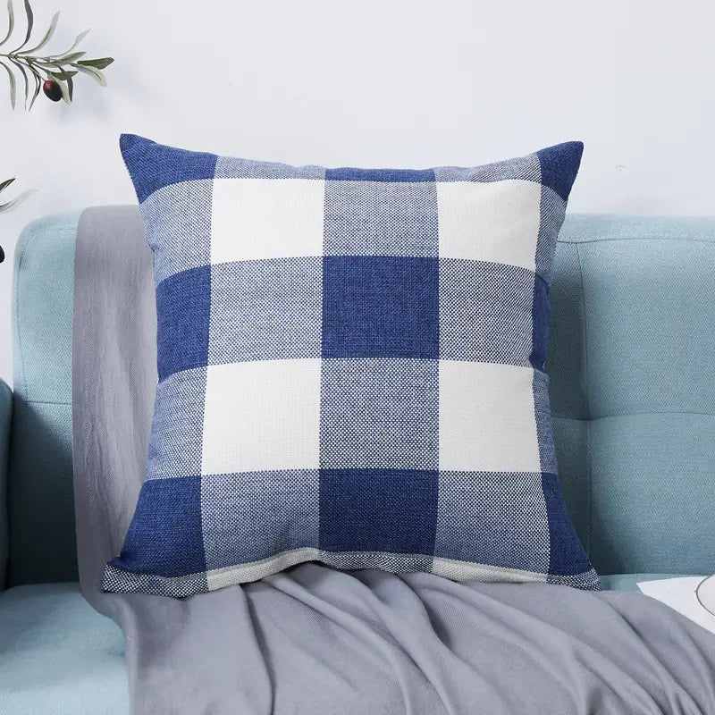 A square throw pillow with a classic large-scale gingham plaid pattern in navy blue and white. The pillow features a textured, linen-like fabric and is shown resting on a light blue sofa next to a grey draped blanket, creating a cozy and traditional aesthetic.