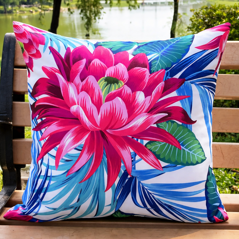 Outdoor 18x18 pillow cover featuring a large pink tropical flower with blue and green leaves on a white background, displayed on a wooden park bench near a lake. Waterproof cover, insert not included.