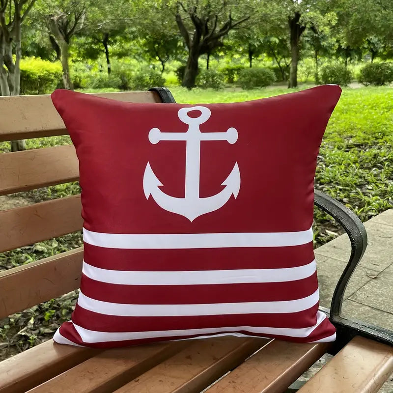 Vibrant red outdoor pillow cover featuring a bold white anchor silhouette on the top half and classic navy-style horizontal white stripes on the bottom half, displayed on an outdoor wooden park bench for nautical décor - 18x18.