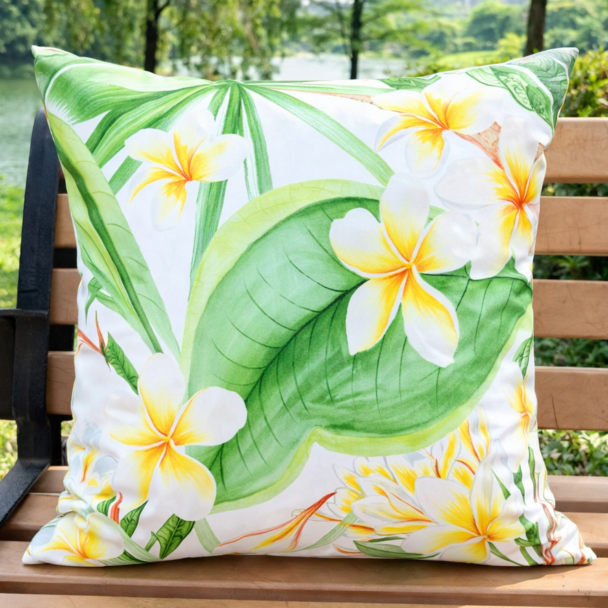 White outdoor pillow cover featuring yellow and white plumeria flowers with green leaves, displayed on a wooden bench in a garden setting. Waterproof 18x18 cover, insert not included.